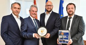 Vier Männer stehen mit einem gewonnenen Preis sowie einem Tablet vor Fahnen