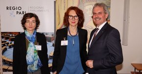 Zwei Damen und ein Herr vor einem Plakat mit der Aufschrift "REGIO PARL"